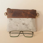 Brown leather case with a gold clip, white notebook, and green-framed glasses on a light gray background