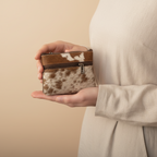 Person holding a small brown and white cowhide wallet against a beige background