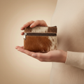 Person holding a small brown and white cowhide leather pouch with a zipper on a beige background.