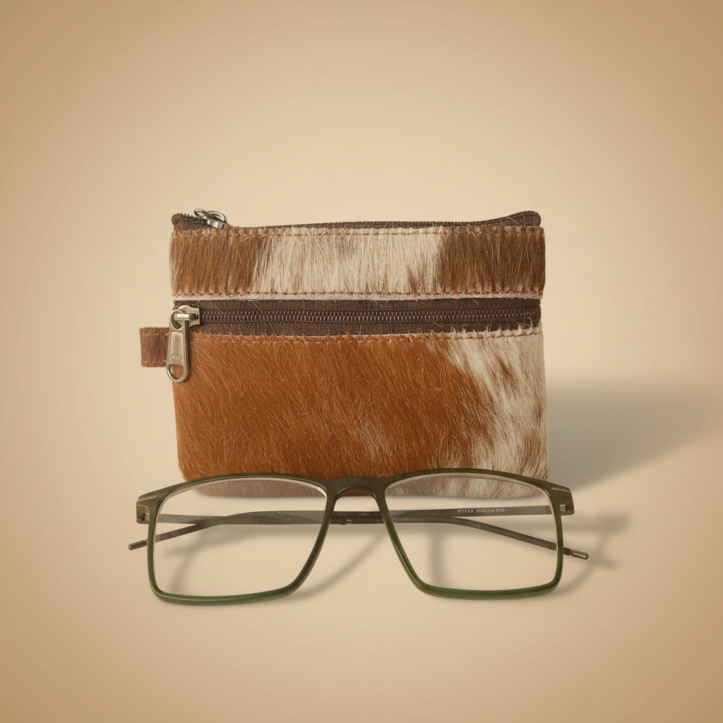 Brown and white cowhide pouch with a zipper and a pair of glasses on a light gray background