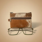 Brown and white cowhide pouch with a zipper and a pair of glasses on a light gray background