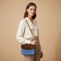 Woman holding a crossbody bag with leopard print and blue section against a beige background