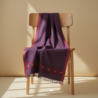 Purple blanket with red border draped over a wooden chair against a beige wall.